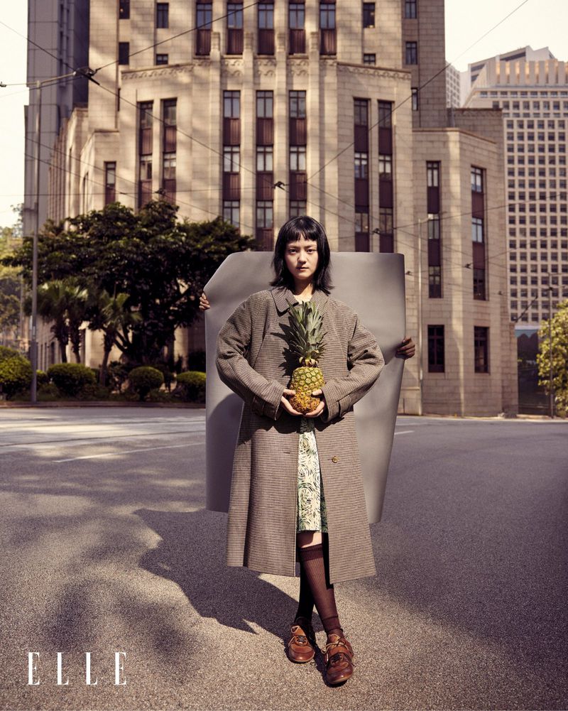 Young woman stands on a city street holding a pineapple, wearing a long coat and patterned dress with a large building in background.