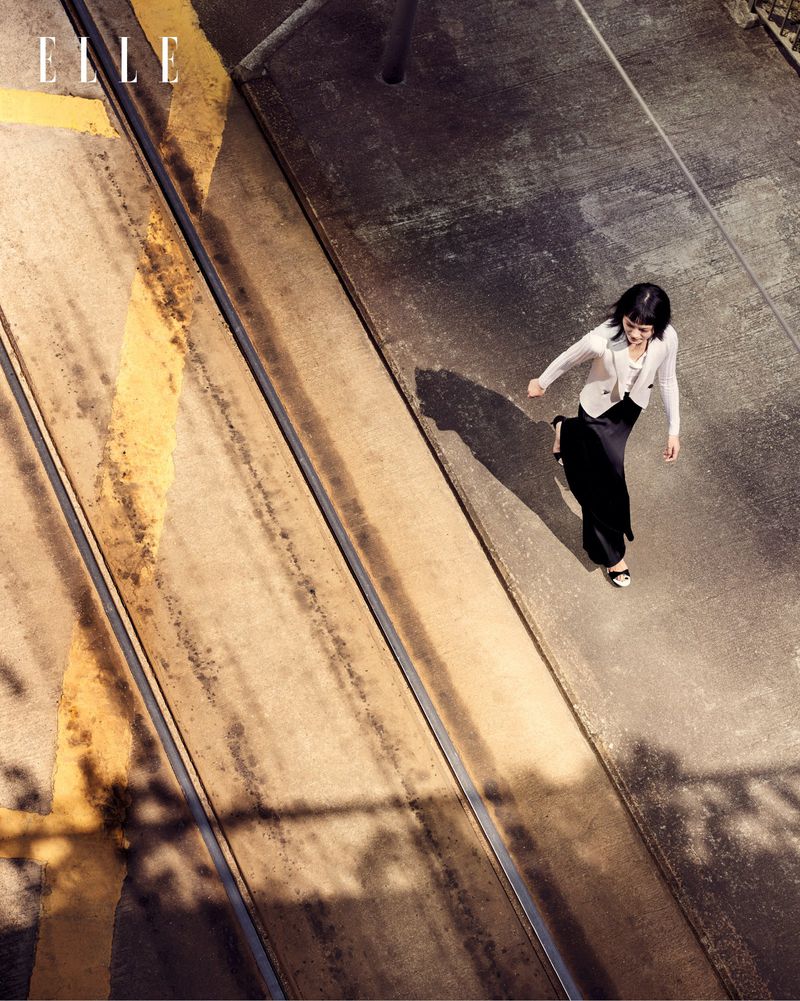 Overhead shot of a woman in a black dress and white blazer walking on a patterned concrete surface with rail tracks in a fashion editorial.
