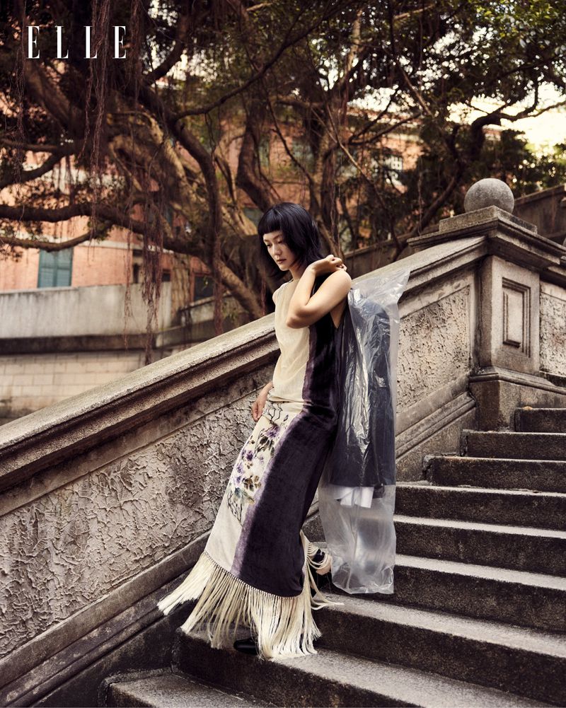 A fashionable woman poses on stone stairs outdoors, holding a garment bag in a street style fashion editorial for Elle magazine.