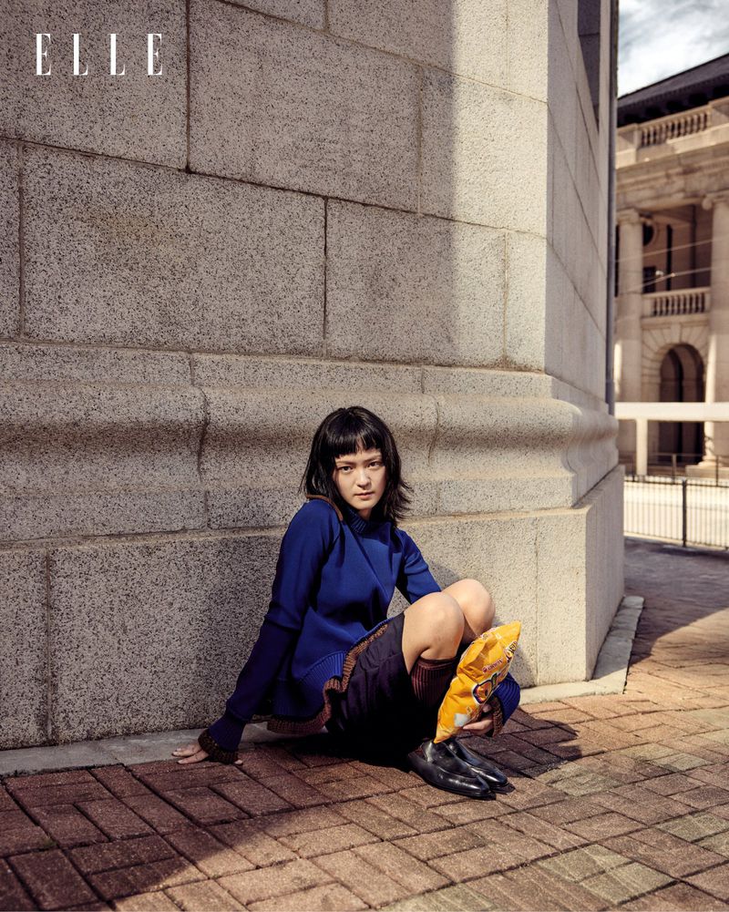 Stylish woman in blue dress sits on brick ground next to building, with Elle magazine logo visible in the upper left corner.