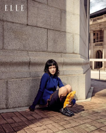 Stylish woman in blue dress sits on brick ground next to building, with Elle magazine logo visible in the upper left corner.