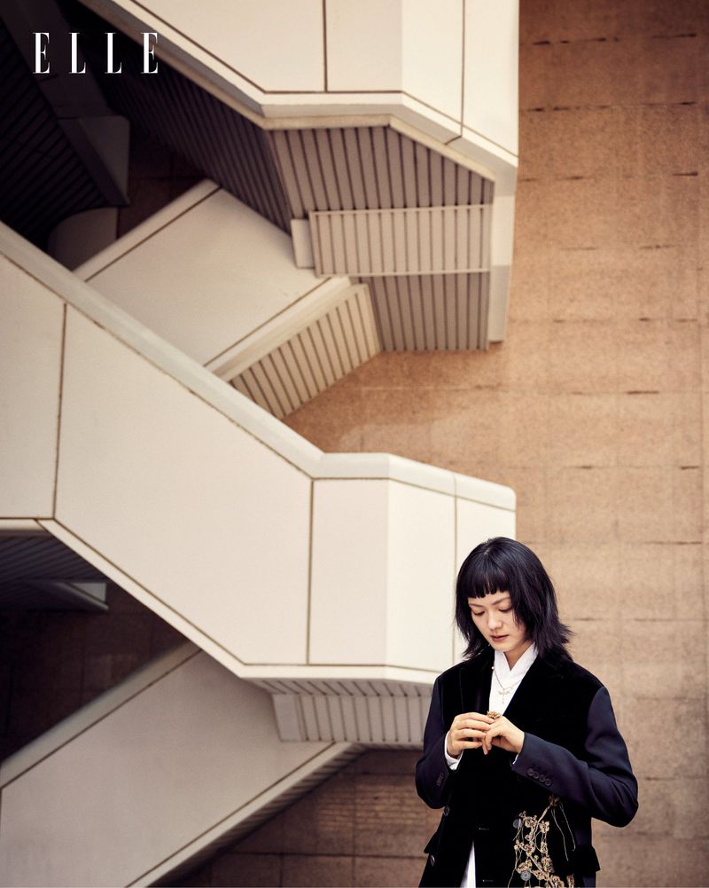 Fashion editorial shot of a woman in a dark blazer standing near a white staircase against a tan textured wall. Elle logo visible.