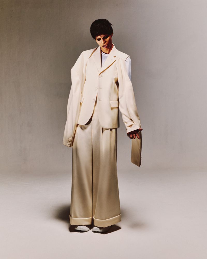 A woman in an oversized ivory suit poses in a photo studio with a serious expression, looking downwards and holding a clutch.