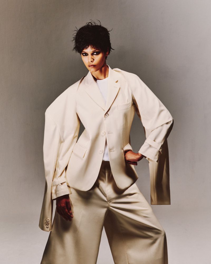 Fashionable woman in oversized beige suit poses in a studio. The suit jacket is draped on her shoulders, adding to the avant-garde look.
