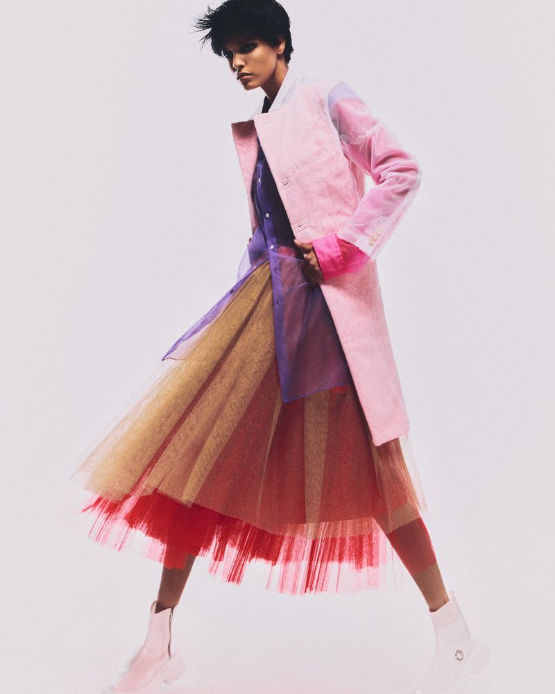 Fashionable person strikes a pose wearing a pink coat, colourful tutu skirt, and white boots in a studio setting with a white backdrop.
