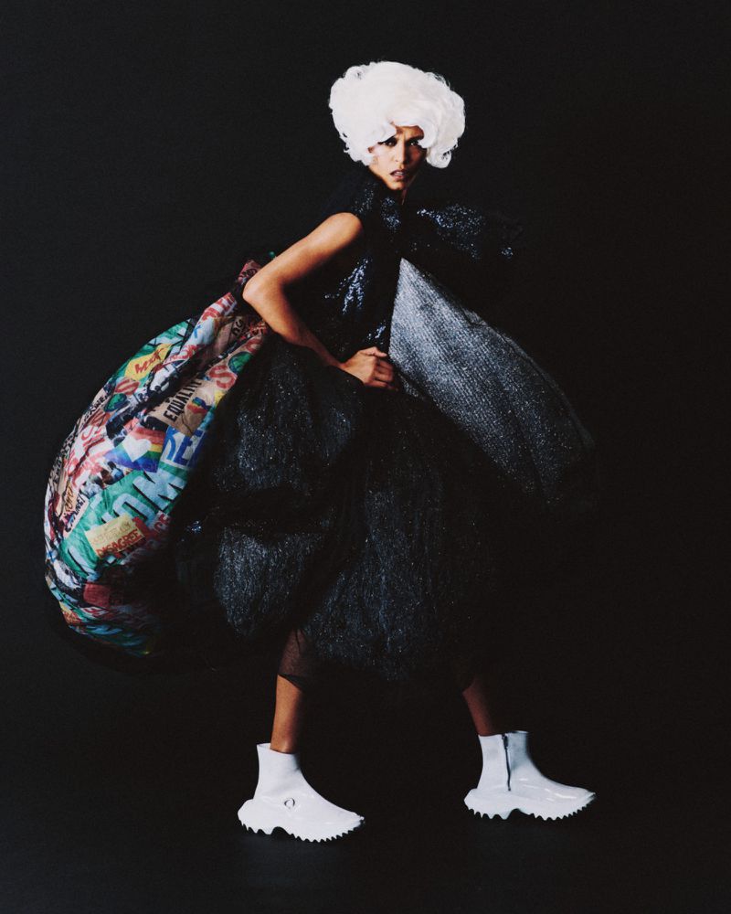 Fashion editorial shot of a woman in a black dress with a large white wig posing in a dark studio with colored fabric detail.