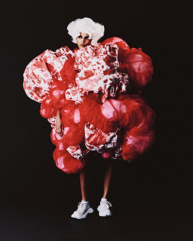 Fashion shot of a woman in an oversized red and white dress with puffy details, accessorized with white sneakers against a dark backdrop.