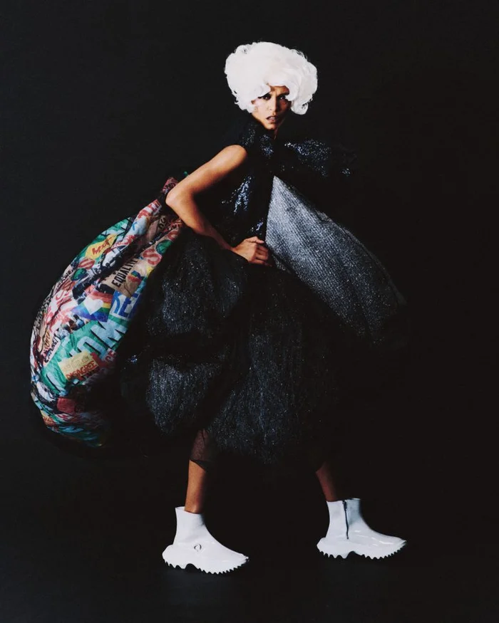 Model wearing a voluminous dark textured outfit with a large colorful patterned bustle and white wig against a black background