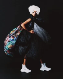 Model wearing a voluminous dark textured outfit with a large colorful patterned bustle and white wig against a black background