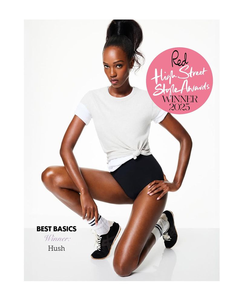 A tanned fashion model poses on her knees in a photo studio wearing a white T-shirt, black bottoms, socks and sneakers against a white backdrop.