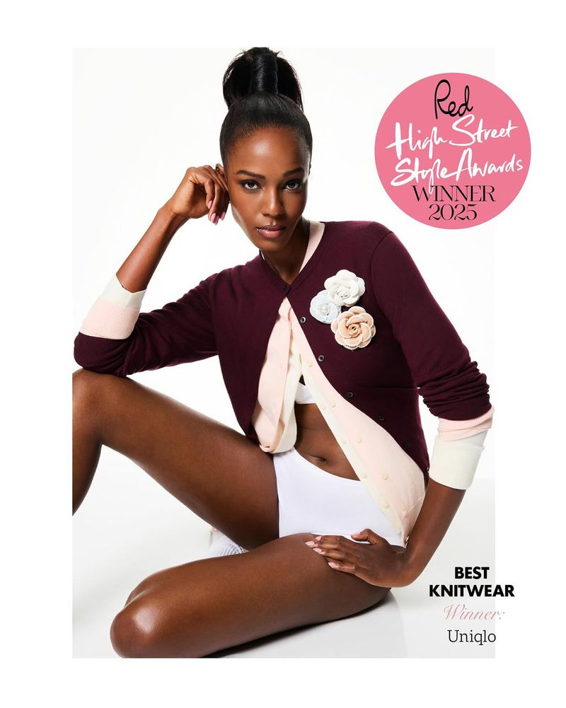A dark-skinned woman poses in a burgundy cardigan, accessorized with floral embellishments, against a clean white backdrop. Fashion shot.