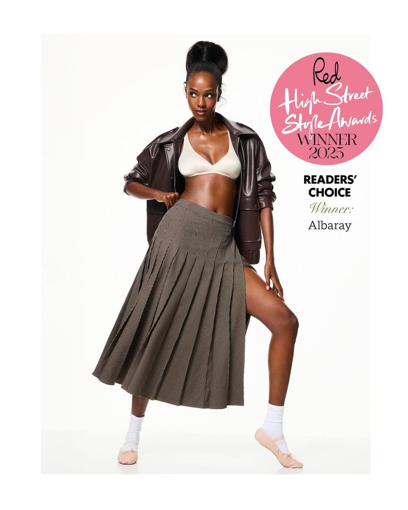 Full shot of a Black female model in a brown jacket, bra top, and plaid skirt, posed against a white backdrop, holding a magazine award.