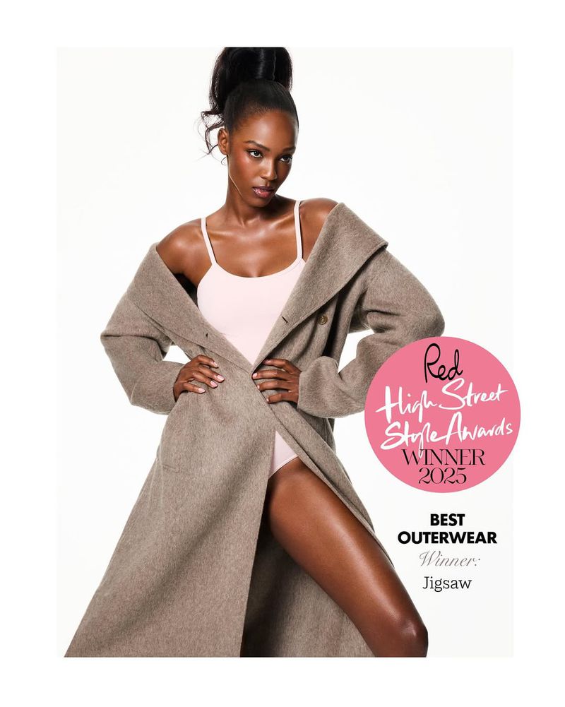 Fashion editorial shot of an adult woman in a long coat, standing indoors with bright lighting, showcasing a stylish and elegant look.