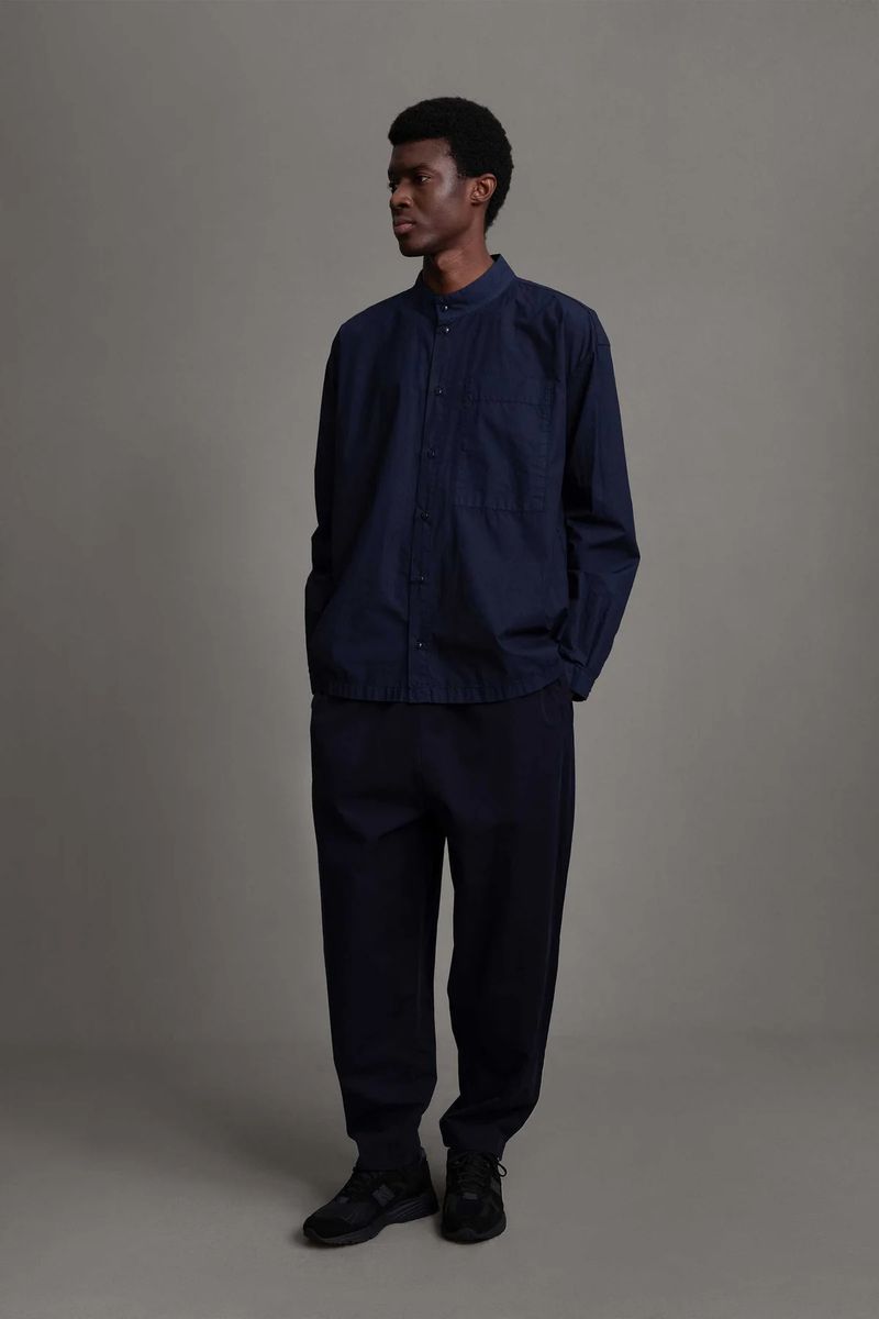 Full body shot of an adult man in a blue button-down shirt and dark pants, standing in a neutral studio with a soft and diffused light.