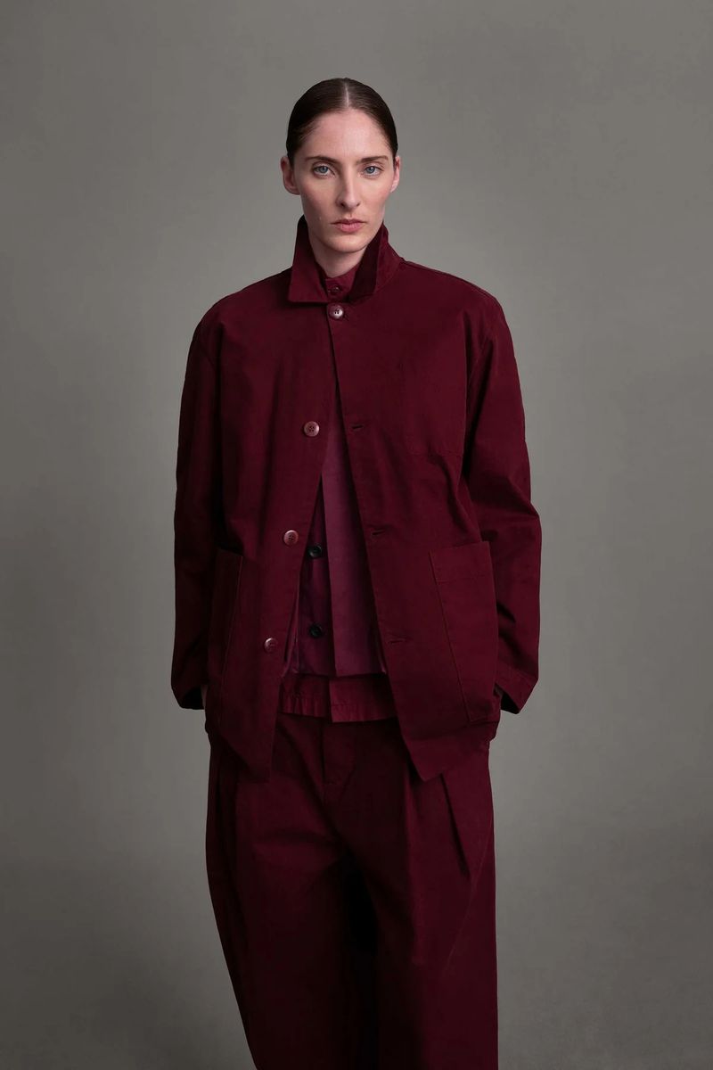 Woman in monochrome burgundy fashion ensemble with structured jacket, button-down shirt, and wide-leg trousers against a grey backdrop.