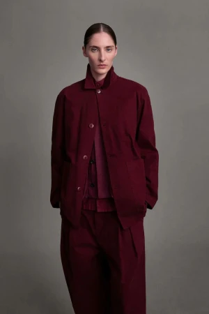 Woman in monochrome burgundy fashion ensemble with structured jacket, button-down shirt, and wide-leg trousers against a grey backdrop.