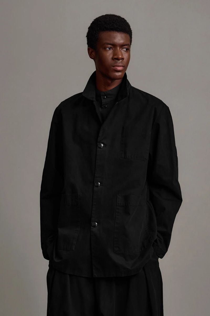 Black male model in a black collared shirt and open jacket poses in front of a neutral background in a studio portrait.