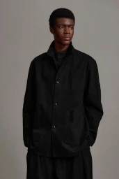 Model Jefferson Obuseri poses in an oversized black buttoned workwear jacket against a neutral grey studio backdrop.