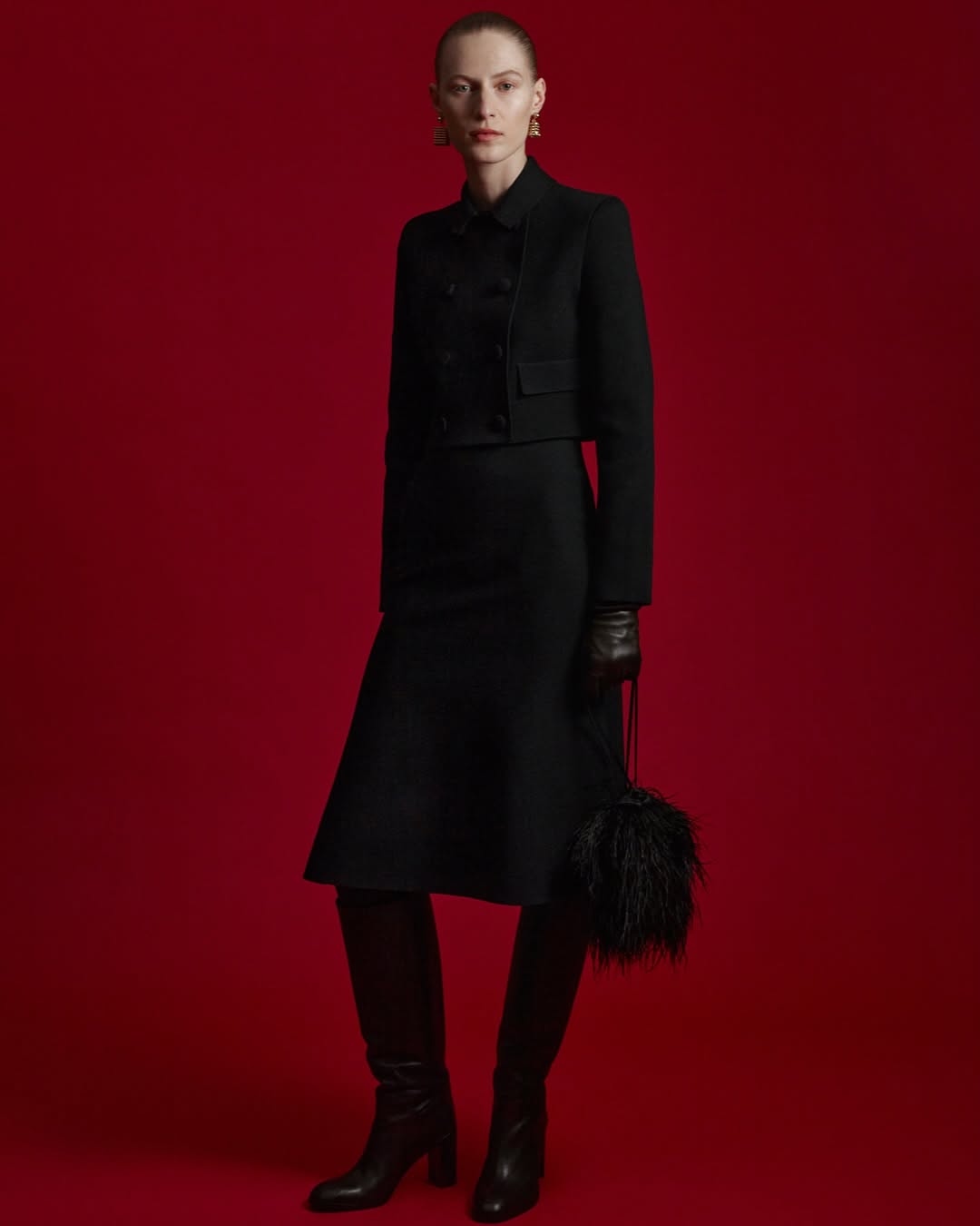 A single woman posing in a black skirt suit, boots, and gold earrings against a dark red background in a studio shot.