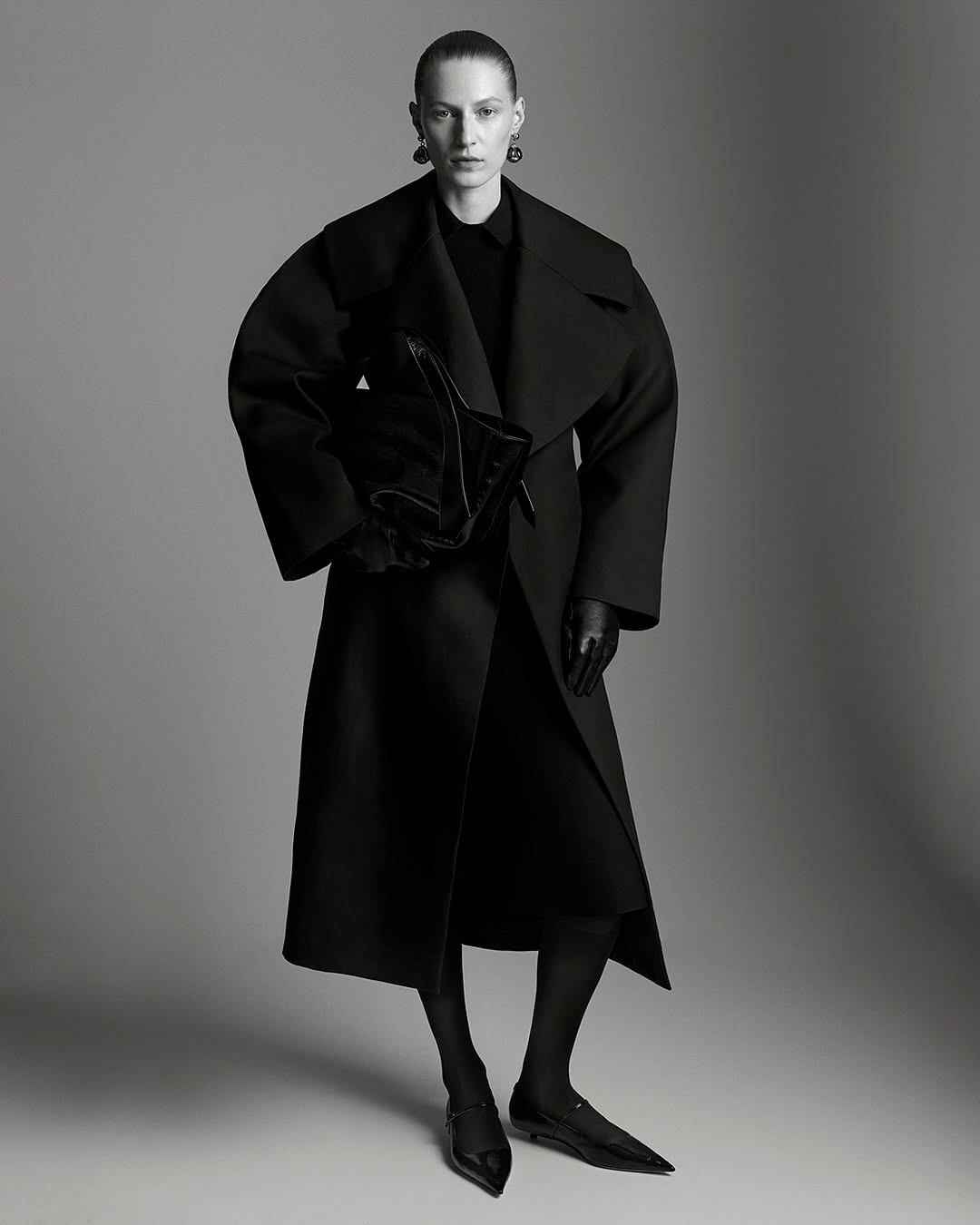 Monochromatic fashion portrait of a woman in a black coat, black dress, and black flats, holding a black purse against a grey backdrop.