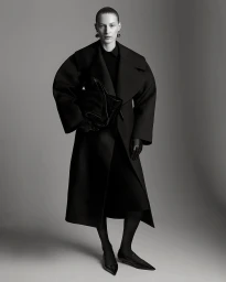 Model in a black coat holding a large clutch bag against a grey seamless studio background.