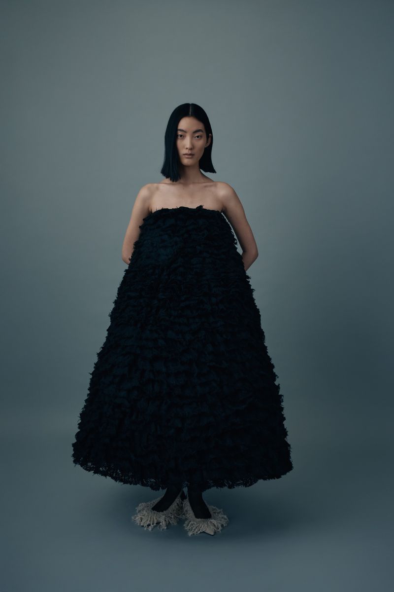 Elegant Asian woman in a large black textured dress posing against a gray backdrop in a fashion editorial studio shot.