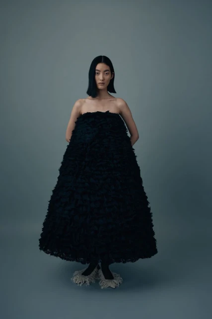 Elegant Asian woman in a large black textured dress posing against a gray backdrop in a fashion editorial studio shot.