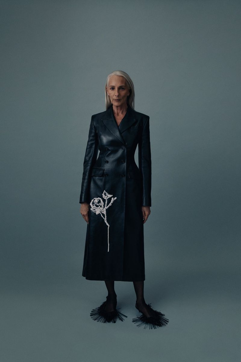Elegant elderly woman in a black double-breasted leather coat with white rose design, posing against a gray studio backdrop.