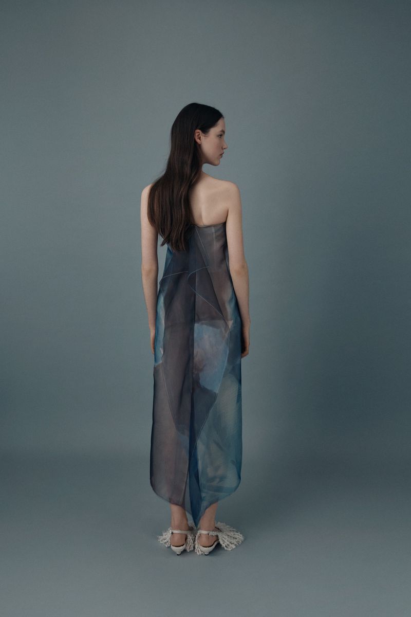 Model viewed from behind in a strapless blue and grey abstract-print midi dress and fringe-detailed heels against a muted blue studio backdrop.