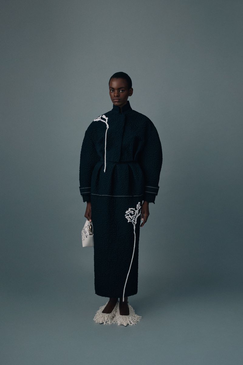Full-length studio shot of a black female model in a dark green dress and coat with white embroidery, holding a small white purse.
