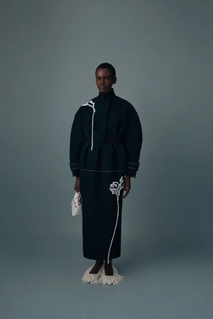 Full-length studio shot of a black female model in a dark green dress and coat with white embroidery, holding a small white purse.