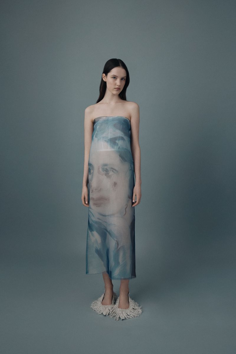 A slender model poses in a blue-tinted sheer strapless dress with a blurred face print, standing against a solid gray background in studio lighting.