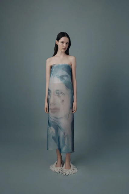 A slender model poses in a blue-tinted sheer strapless dress with a blurred face print, standing against a solid gray background in studio lighting.