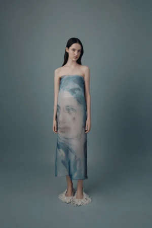 A slender model poses in a blue-tinted sheer strapless dress with a blurred face print, standing against a solid gray background in studio lighting.