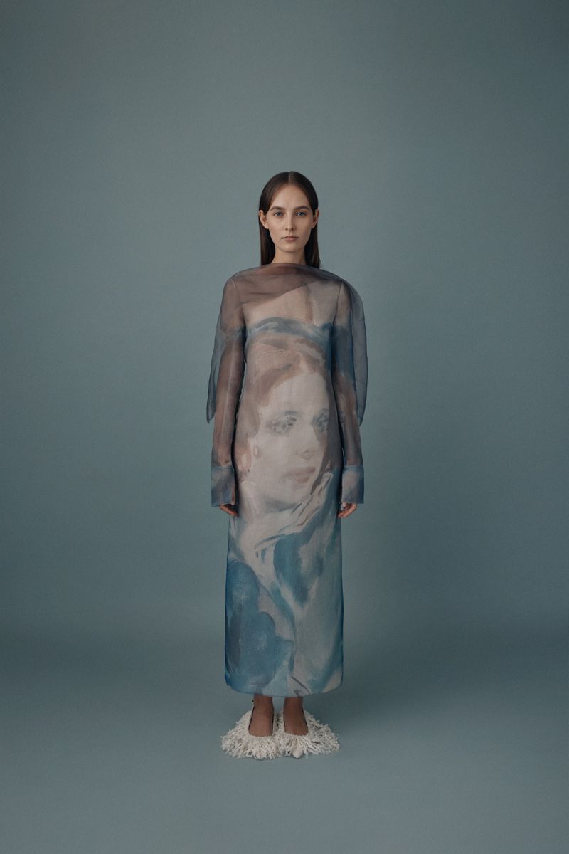Full-length shot of a slender woman with long brown hair wearing a printed sheer dress and cream-colored fuzzy slippers, against a gray background.