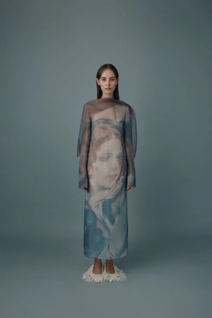 Full-length shot of a slender woman with long brown hair wearing a printed sheer dress and cream-colored fuzzy slippers, against a gray background.