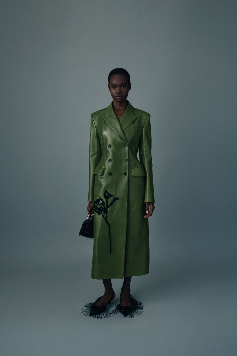 A slender Black woman poses indoors wearing a green double-breasted leather coat, black shoes with feather details, and holding a black purse.