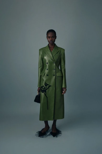 A slender Black woman poses indoors wearing a green double-breasted leather coat, black shoes with feather details, and holding a black purse.