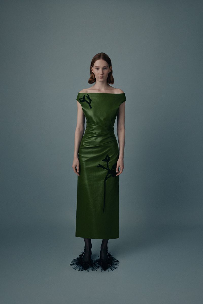 Full-length shot of a fair-skinned fashion model wearing a green off-shoulder dress with black embellishments and textured black heels on gray.