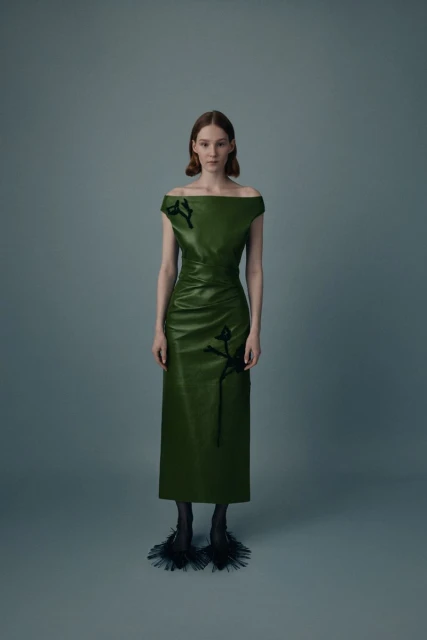 Full-length shot of a fair-skinned fashion model wearing a green off-shoulder dress with black embellishments and textured black heels on gray.