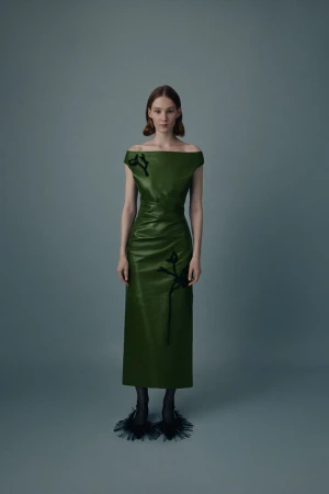 Full-length shot of a fair-skinned fashion model wearing a green off-shoulder dress with black embellishments and textured black heels on gray.