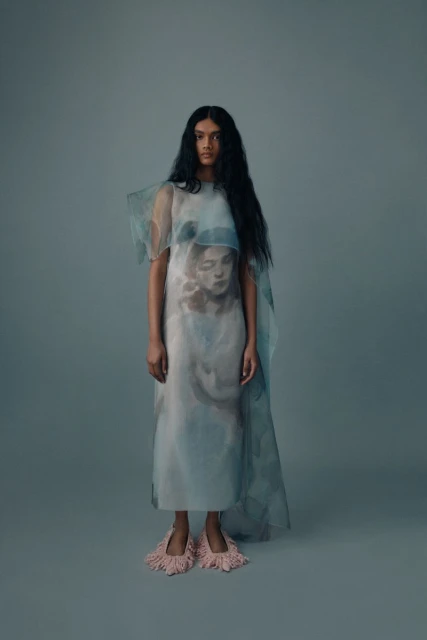 Fashion portrait of an adult female wearing a translucent dress with an image print, posing in front of a grey backdrop.