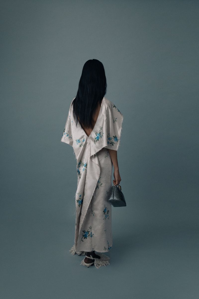 Back view of woman with long dark hair wearing a floral dress and holding a metallic handbag against a grey studio background.