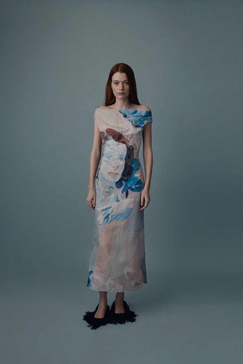 A slender woman with long red hair poses in a painterly blue-and-beige one-shoulder dress and black shoes against a grey background.