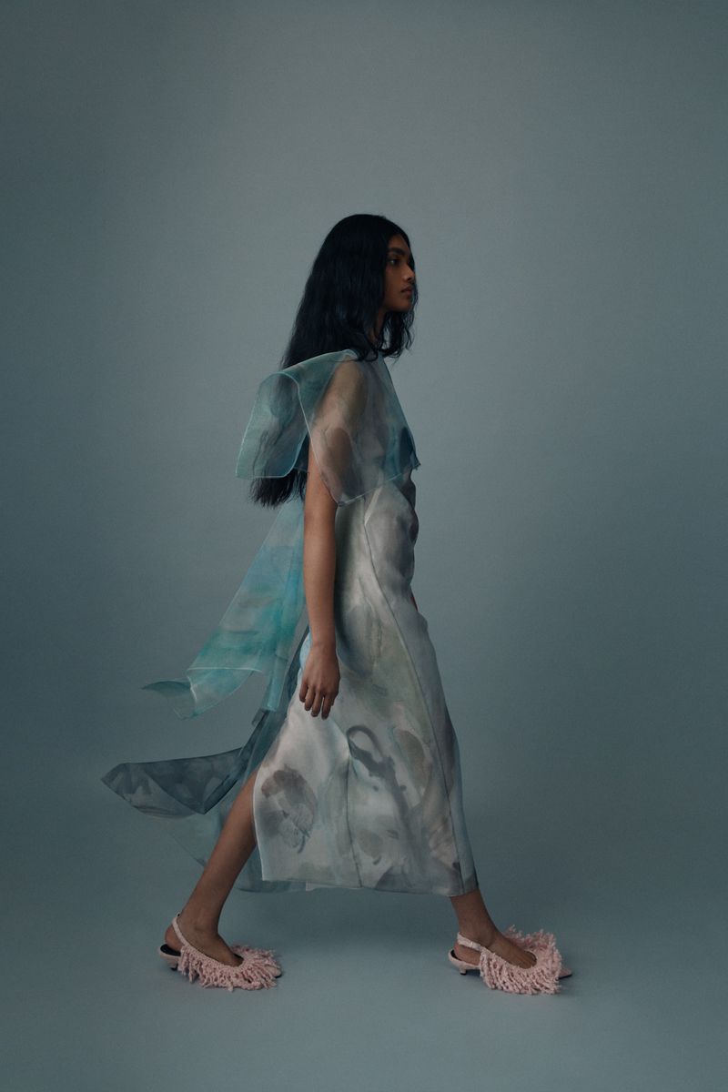 A person with long dark hair wears a light blue and white dress and pink fringe shoes, walking in a brightly lit photo studio.