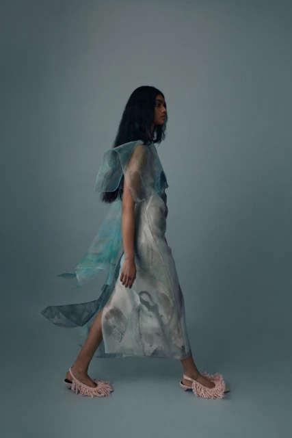 A person with long dark hair wears a light blue and white dress and pink fringe shoes, walking in a brightly lit photo studio.