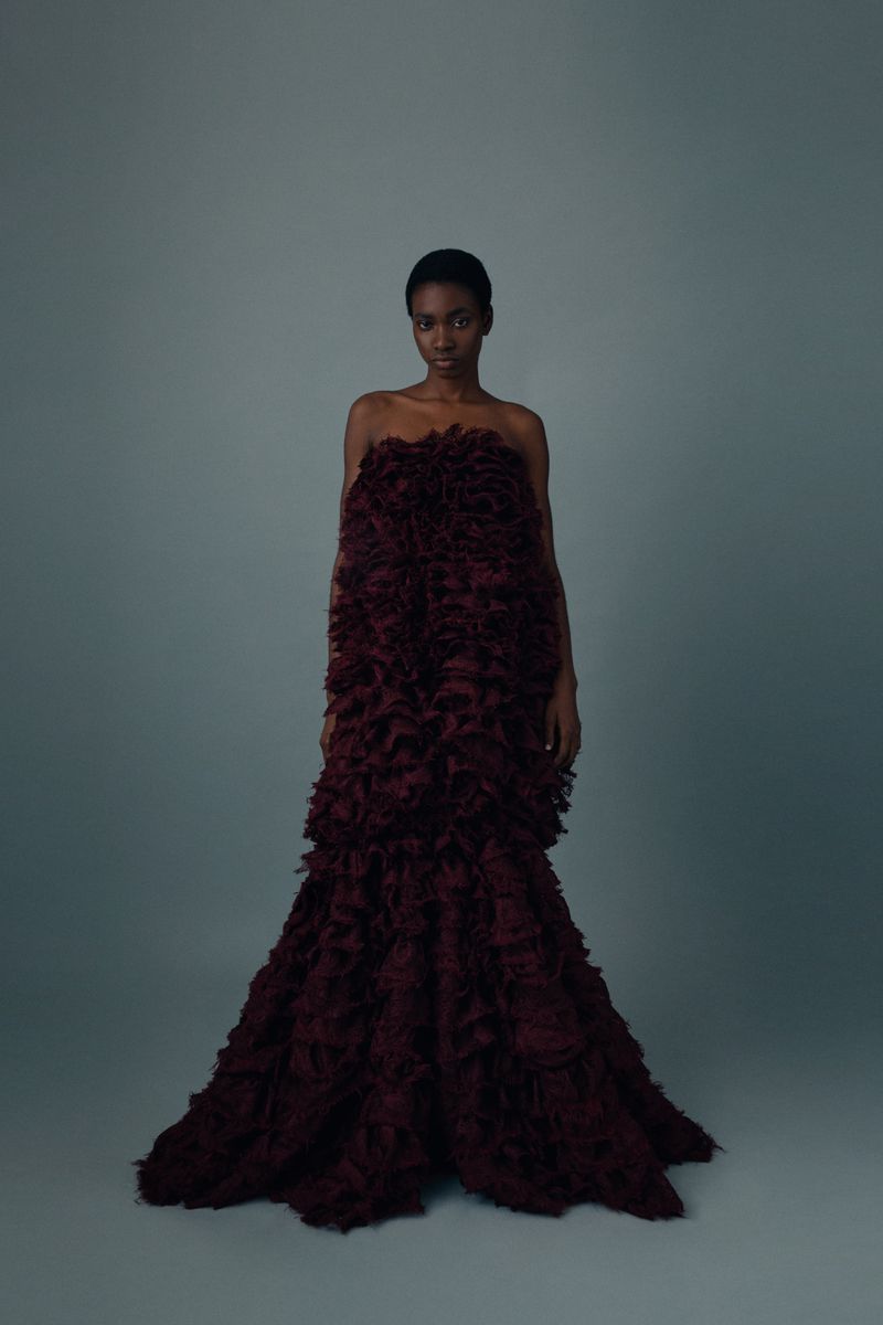 An elegant, African-descent female fashion model poses in a tiered, floor-length dark-red feathery gown against a neutral grey backdrop.