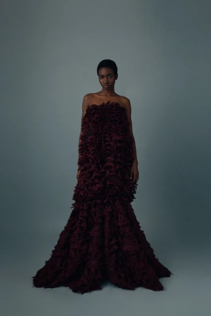 An elegant, African-descent female fashion model poses in a tiered, floor-length dark-red feathery gown against a neutral grey backdrop.