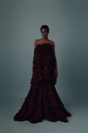 An elegant, African-descent female fashion model poses in a tiered, floor-length dark-red feathery gown against a neutral grey backdrop.
