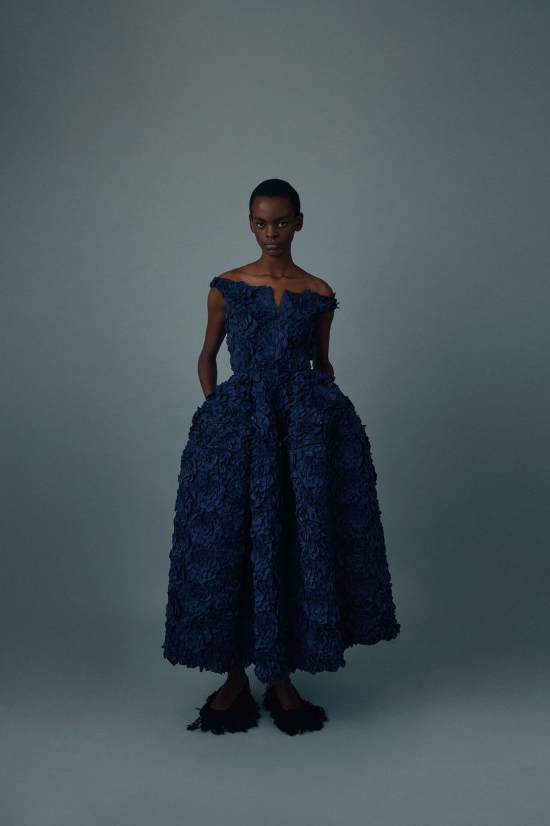 Elegant woman wearing a dark blue, off-the-shoulder textured gown standing indoors against a gray backdrop in a studio shot.